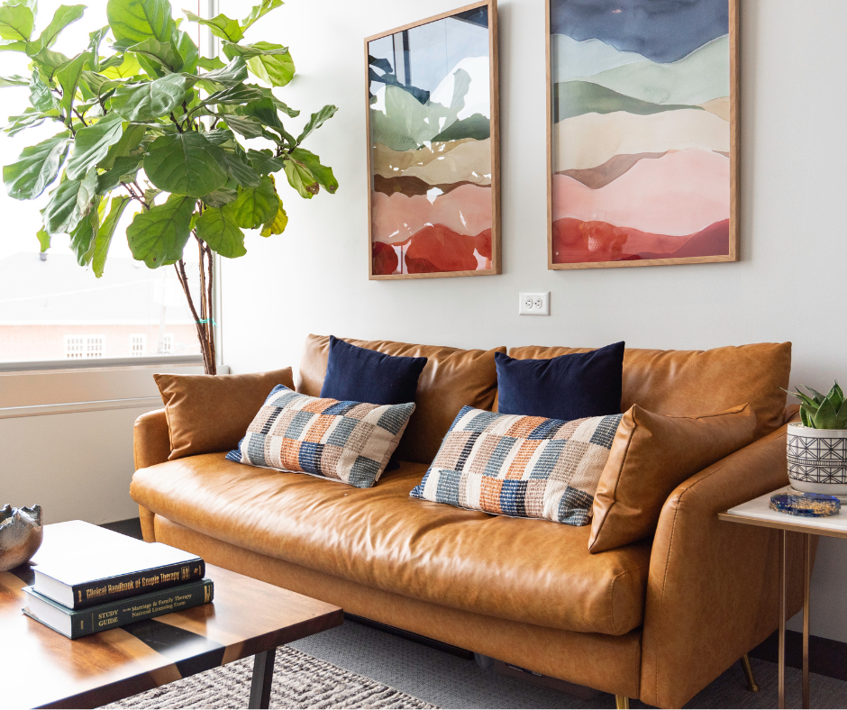 An interior view of our Fort Worth, TX counseling space, featuring a tan leather sofa and books titled 'Anxiety Isn't Always Obvious,' with a pillow labeled 'Fort Wellness Counseling in Fort Worth, TX' and an abstract landscape print.